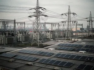 Overbelaste hoogspanningslijnen en transformatoren boven een industriële haven in Rotterdam-stijl, met zonnepanelen op magazijndaken.